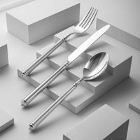 A fork, knife, and spoon from Ricci's Solitaire Flatware 5 Piece Setting, crafted in 18/10 stainless steel with textured handles, are displayed on white geometric steps to highlight their modern elegance.