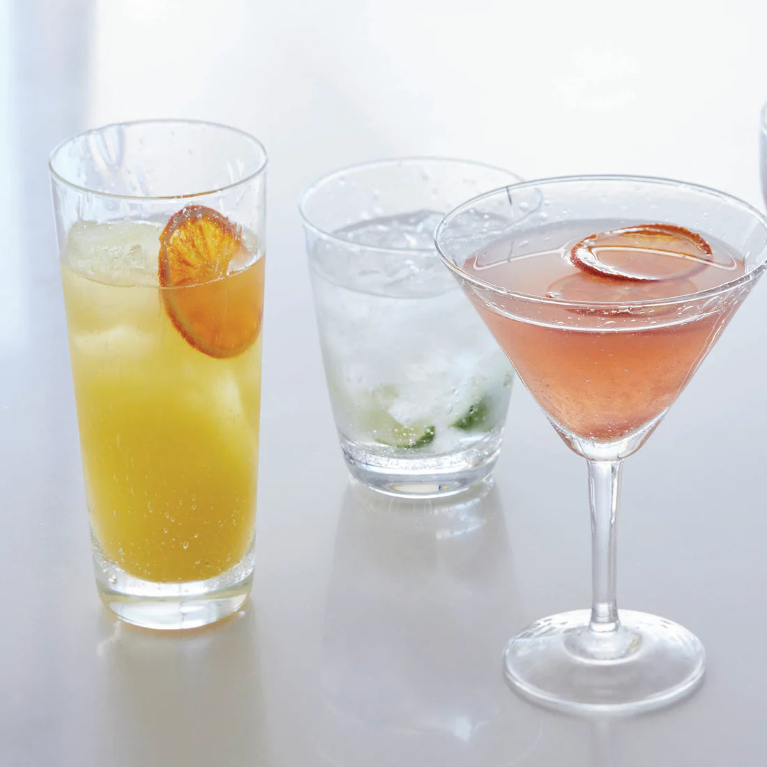 Three cocktails in elegant Mariposa Bellini Glassware: a yellow drink with orange slice in a tall glass, a clear drink with mint in a stemless wine glass, and a pink drink with orange slice in a martini glass.