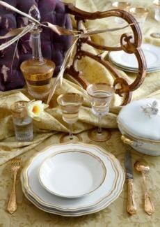 Elegant table setting with Raynaud Polka Gold Dinnerware, gold flatware, wine glasses, a decanter, covered dish, and a yellow flower on a gold-patterned tablecloth beside a wooden chair.