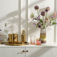 A bar setup with a gold tray, ice bucket, and cocktail shaker sits on a white cabinet. Bottles, two mixed drinks in Aaron Pink Glassware by Blue Pheasant, and a vase with purple and pink flowers are displayed against a white wall.