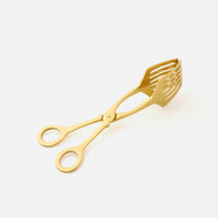 The Zena Serving Tongs by Blue Pheasant are gold-colored brass utensils with slotted, scoop-like ends and finger loops, designed for serving food. They are elegantly displayed on a plain white background.