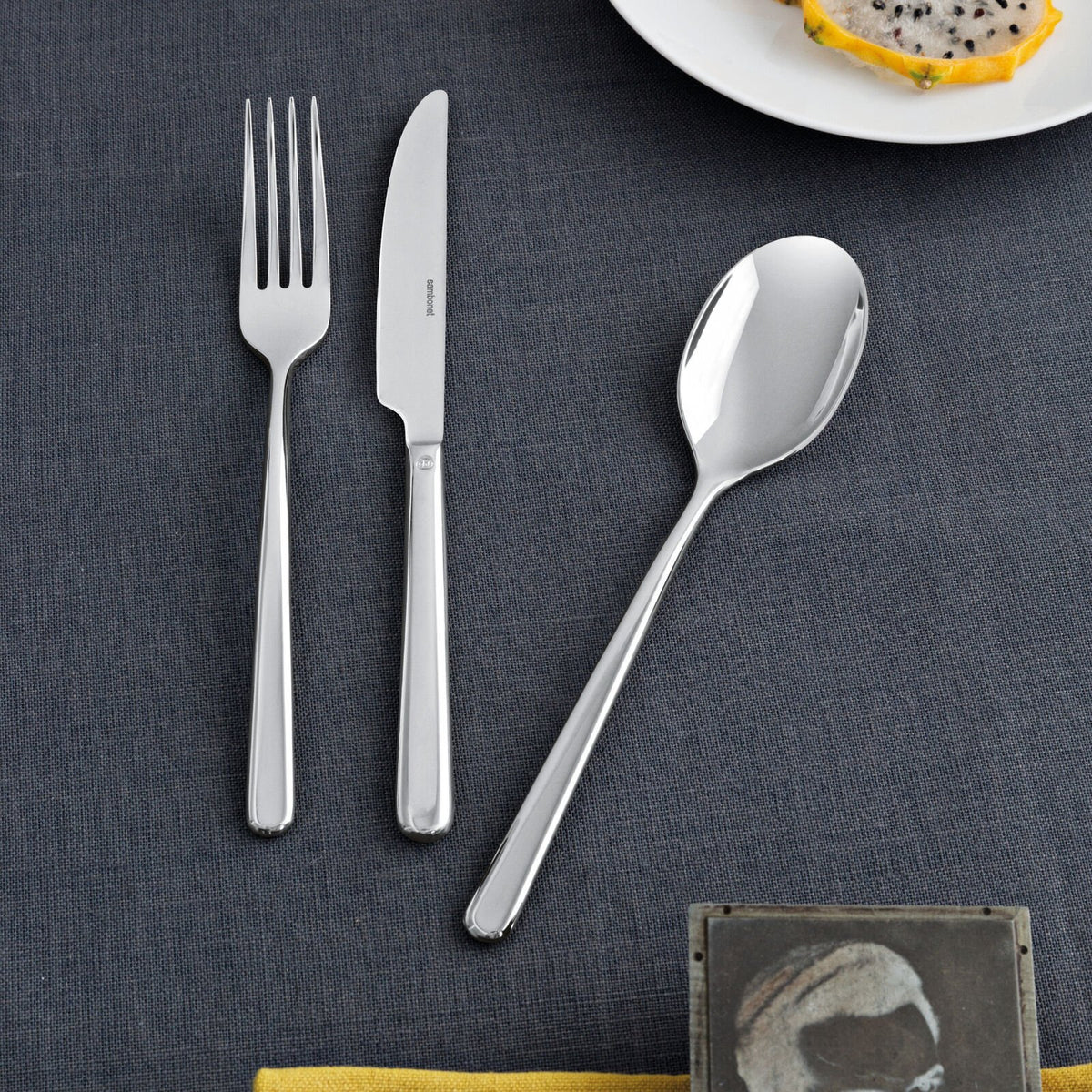 The Sambonet Linear Solid Handle Flatware 5 Place Setting is neatly arranged on a dark gray tablecloth, alongside a white plate with dragon fruit slices and the corner of a yellow napkin.