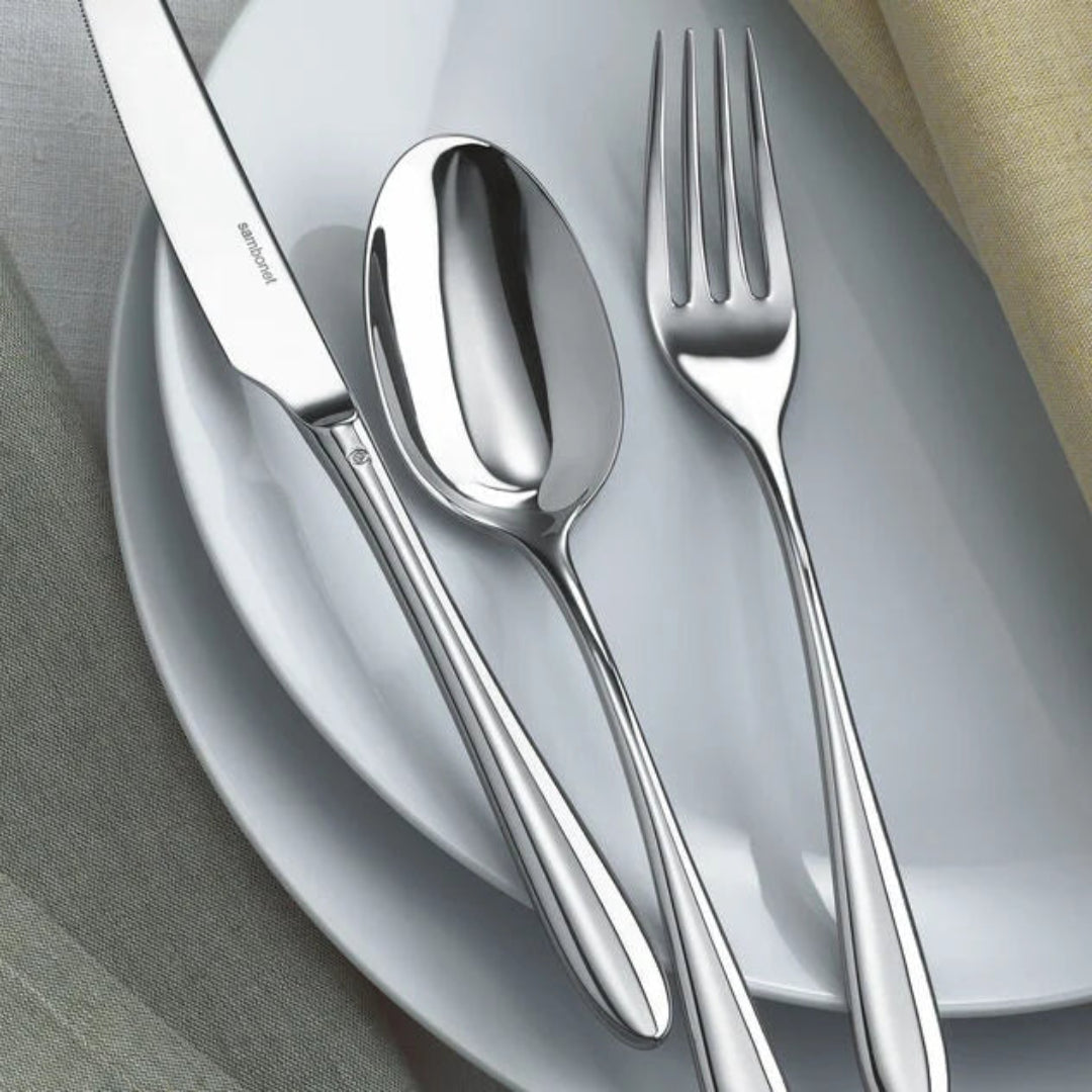 The Sambonet Dream Solid Handle Flatware 5 Piece Setting, featuring a knife, spoon, and fork, sits neatly on stacked white plates with light gray and yellow napkins placed nearby.