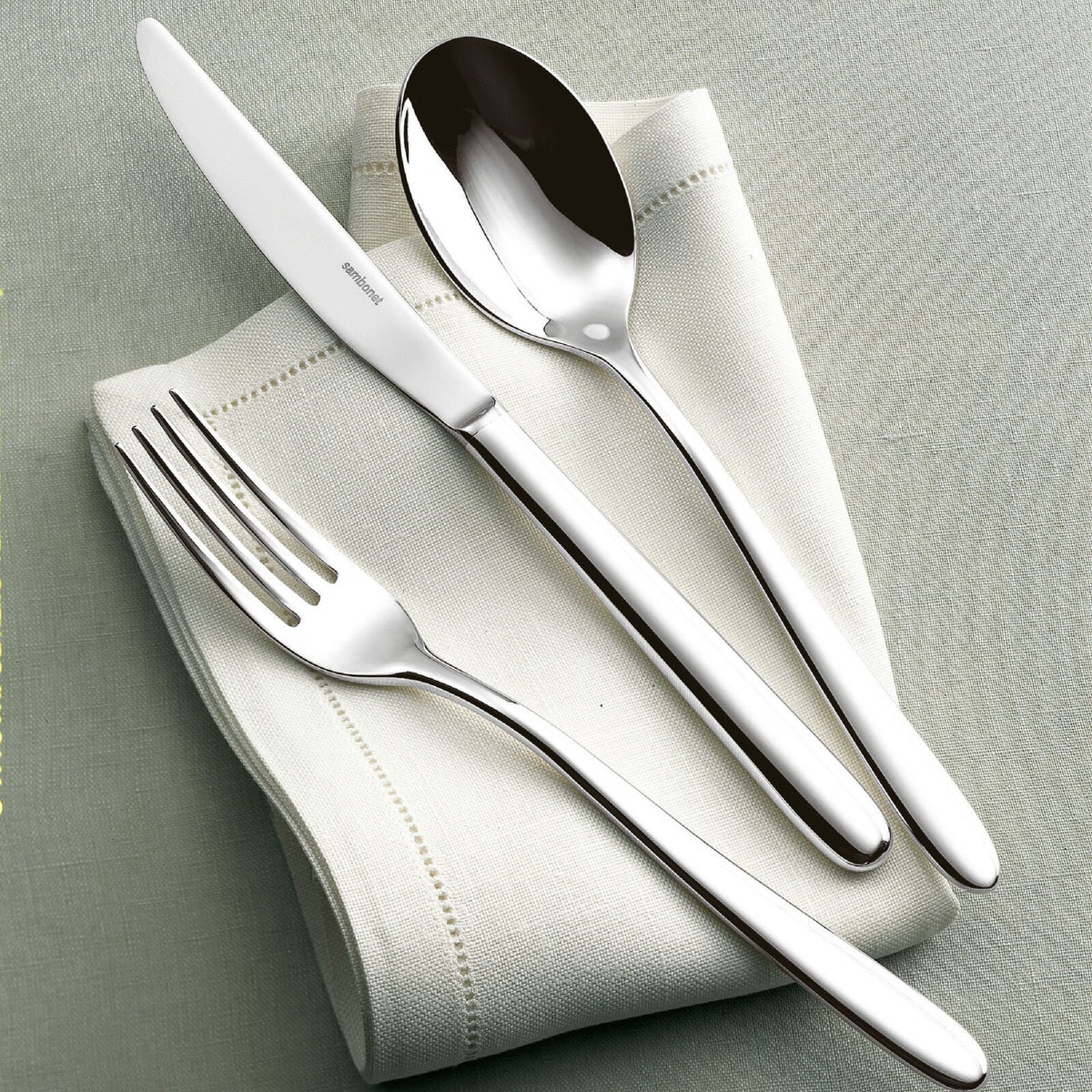 The Sambonet Hannah Flatware 5 Piece Setting in stainless steel is elegantly displayed on a folded white cloth napkin atop a pale green tablecloth.