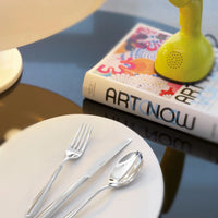 A white plate with the Sambonet Hannah Flatware 5 Piece Setting in stainless steel is on a reflective table beside a colorful ART NOW book and a small yellow lamp.