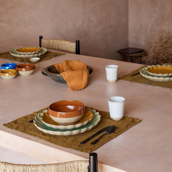 A table set for four features Costa Nova’s Marrakesh Dinnerware Orange—Moroccan-inspired plates, bowls, napkins, and cups on tan placemats. The recycled clay complements neutral tones and textured walls with a wooden stool in the background.