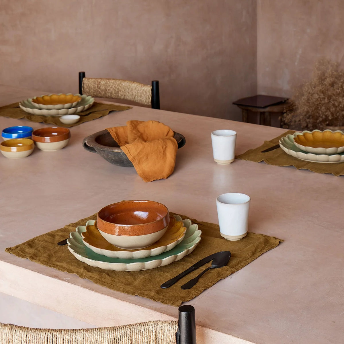 A dining table set with vibrant Casafina/Costa Nova Marrakesh Dinnerware Yellow ceramic plates and eco-stoneware bowls, paired with cloth napkins, cutlery, and white cups atop brown placemats. Four place settings are arranged in a minimalist, earthy-toned room.