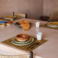 A dining table set with vibrant Casafina/Costa Nova Marrakesh Dinnerware Yellow ceramic plates and eco-stoneware bowls, paired with cloth napkins, cutlery, and white cups atop brown placemats. Four place settings are arranged in a minimalist, earthy-toned room.