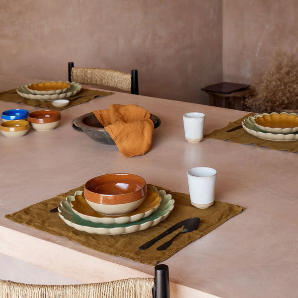 A dining table set with vibrant Casafina/Costa Nova Marrakesh Dinnerware Yellow ceramic plates and eco-stoneware bowls, paired with cloth napkins, cutlery, and white cups atop brown placemats. Four place settings are arranged in a minimalist, earthy-toned room.