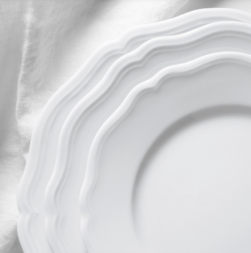 Three overlapping Argent White Dinnerware plates by Raynaud, featuring scalloped edges, rest on crumpled white fabric, creating a soft and elegant monochrome composition.