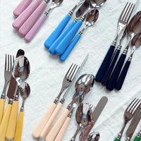 Assorted Capdeco Altea Flatware 5 Piece Setting forks, knives, and spoons with stainless steel heads and pastel or dark blue handles—pink, blue, yellow, cream, and dark blue—are arranged on a white fabric background.