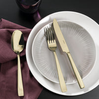 A set table displays stacked white and gray plates, Sambonet’s Flat PVD Flatware 5 Piece Setting in Champagne—a gold knife and fork crossed on top—plus a gold spoon on a mauve napkin, dark glass above, all on a dark tabletop. Made in Italy.