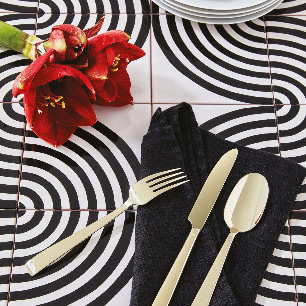 A close-up of a table set with Sambonet's Flat PVD Flatware 5 Piece Setting in Champagne on a black napkin, red flowers, and a bold black-and-white spiral tiled tabletop. Plates are partially visible at the top.