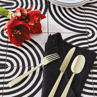 A close-up of a table set with Sambonet's Flat PVD Flatware 5 Piece Setting in Champagne on a black napkin, red flowers, and a bold black-and-white spiral tiled tabletop. Plates are partially visible at the top.