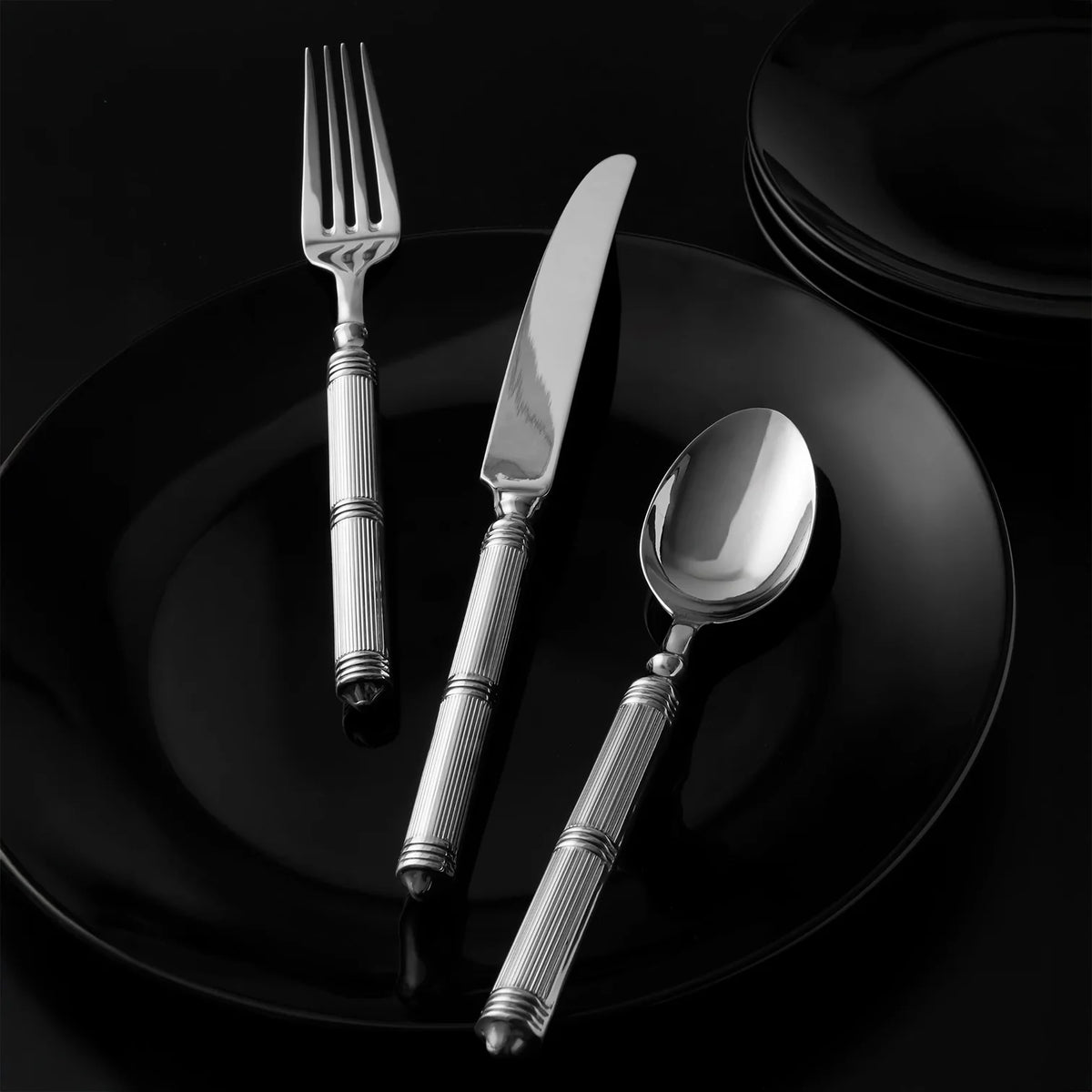The Ricci Castello Flatware Silver 5-Piece Setting, featuring patterned handles and crafted from 18/10 stainless steel, is arranged on a black plate with stacked black plates in the background against a dark surface.