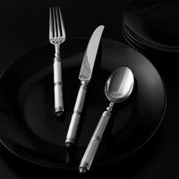 The Ricci Castello Flatware Silver 5-Piece Setting, featuring patterned handles and crafted from 18/10 stainless steel, is arranged on a black plate with stacked black plates in the background against a dark surface.