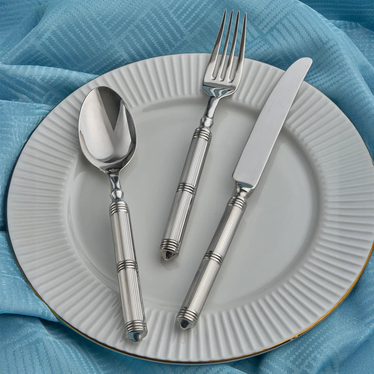 A white plate with a ridged edge sits on a blue textured cloth, holding a silver spoon, fork, and knife from the Ricci Castello Flatware Silver 5-Piece Setting.