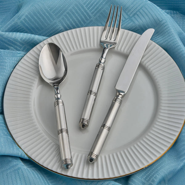 A white plate with a ridged edge sits on a blue textured cloth, holding a silver spoon, fork, and knife from the Ricci Castello Flatware Silver 5-Piece Setting.