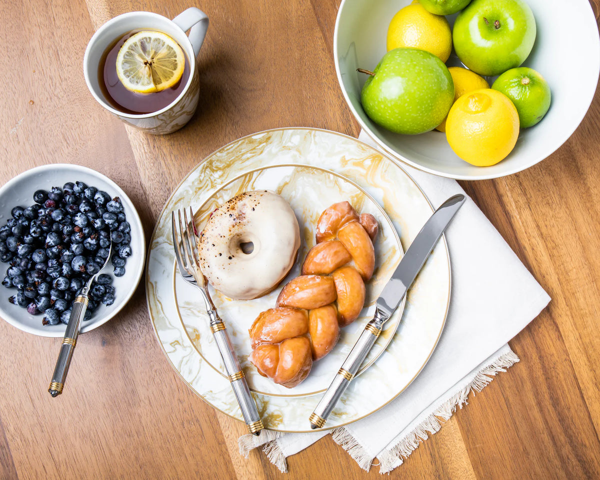 A breakfast scene features the Ricci Castello Flatware Gold 5-Piece Setting beside a cup of tea with lemon, a marble plate with a glazed donut and braided pastry, plus bowls of blueberries, green apples, and lemons on a wooden table.