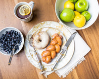 A breakfast scene features the Ricci Castello Flatware Gold 5-Piece Setting beside a cup of tea with lemon, a marble plate with a glazed donut and braided pastry, plus bowls of blueberries, green apples, and lemons on a wooden table.