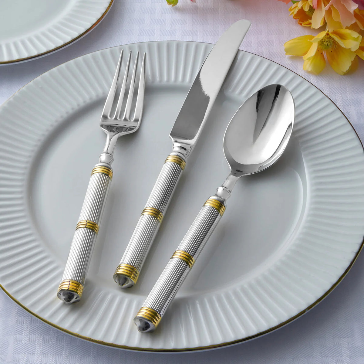 A white, fluted dinner plate with Ricci's Castello Flatware Gold 5-Piece Setting—fork, knife, and spoon with 24kt gold-accented handles—rests on a white tablecloth beside yellow flowers and another plate.