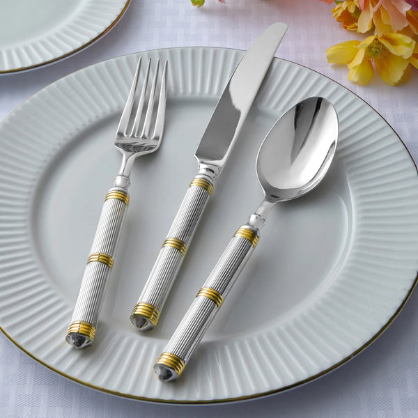 A white, fluted dinner plate with Ricci's Castello Flatware Gold 5-Piece Setting—fork, knife, and spoon with 24kt gold-accented handles—rests on a white tablecloth beside yellow flowers and another plate.