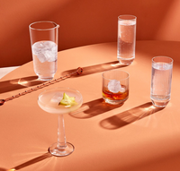 Five cocktails in various glasses, including a set of 2 NUDE Big Top Coupe Glasses by Nude Glassware, sit on a peachy-orange table under bright light with distinct shadows. A cocktail stirrer is nearby. All glassware is crafted from lead-free crystal.