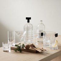 A set of Orrefors Kosta Boda Street Highball Glasses (set of 2) is displayed on a wooden surface with various garnishes and bottles, alongside other elegant crystal glassware including a decanter, martini glass, and tumblers.