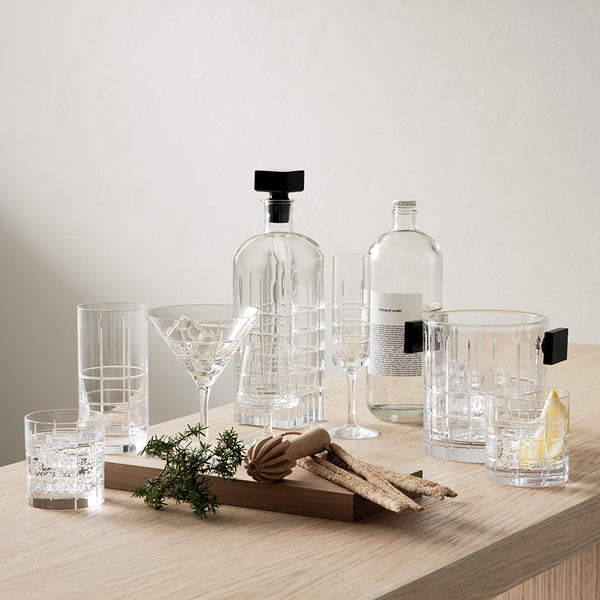 A set of Orrefors Kosta Boda Street Highball Glasses (set of 2) is displayed on a wooden surface with various garnishes and bottles, alongside other elegant crystal glassware including a decanter, martini glass, and tumblers.