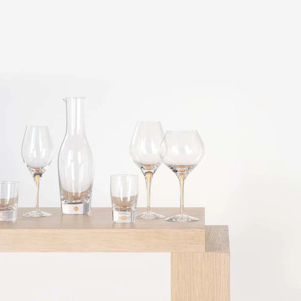 A wooden table displays assorted clear glassware, including the Orrefors Kosta Boda Intermezzo Champagne Glass Gold Set of 2, wine glasses, a carafe, and tumblers, all arranged neatly against a plain white background.