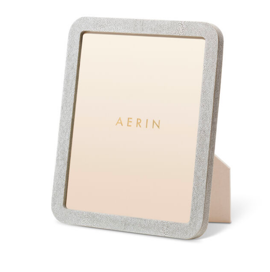 The Aerin Modern Shagreen Frame Dove is a rectangular standing photo frame with a textured, light gray shagreen finish and an easel back. "AERIN" is printed in gold on the beige insert for a contemporary touch.