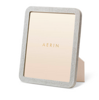The Aerin Modern Shagreen Frame Dove is a rectangular standing photo frame with a textured, light gray shagreen finish and an easel back. "AERIN" is printed in gold on the beige insert for a contemporary touch.