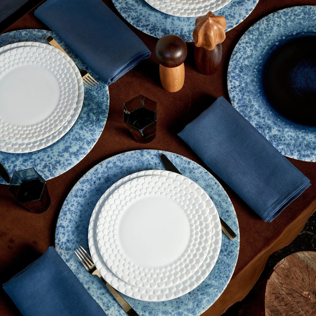 A table set with white textured plates on L'Objet Terra Platter Charger Indigo, navy napkins, black flatware, dark glasses, wooden salt and pepper shakers, and elegant porcelain placemats with cork backing atop a brown tablecloth.
