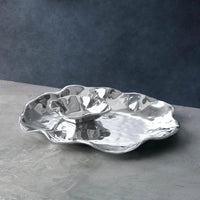 The Beatriz Ball Vento Bergamo Oven Platter & Bowl, reflective with wavy silver edges and crafted from aluminum alloy, sits on a gray tabletop against a dark blue-gray background.