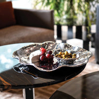 The Beatriz Ball Vento Bergamo Oven Platter & Bowl, a shiny, wavy aluminum alloy piece, holds cheese, grapes, and olives on a black marble table with a sofa and green plants in the background.