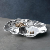The Beatriz Ball Vento Bergamo Oven Platter & Bowl, with wavy edges and a gleaming aluminum alloy finish, holds pistachios on a gray surface against a dark blue background.