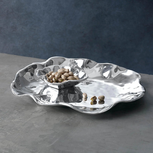 The Beatriz Ball Vento Bergamo Oven Platter & Bowl, with wavy edges and a gleaming aluminum alloy finish, holds pistachios on a gray surface against a dark blue background.