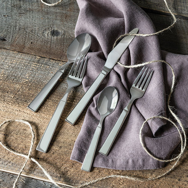 The Simon Pearce Woodstock 5 Piece Setting, modern stainless steel flatware including fork, knife, and spoon, is displayed on a rustic wood surface with a lavender napkin and twine accent.