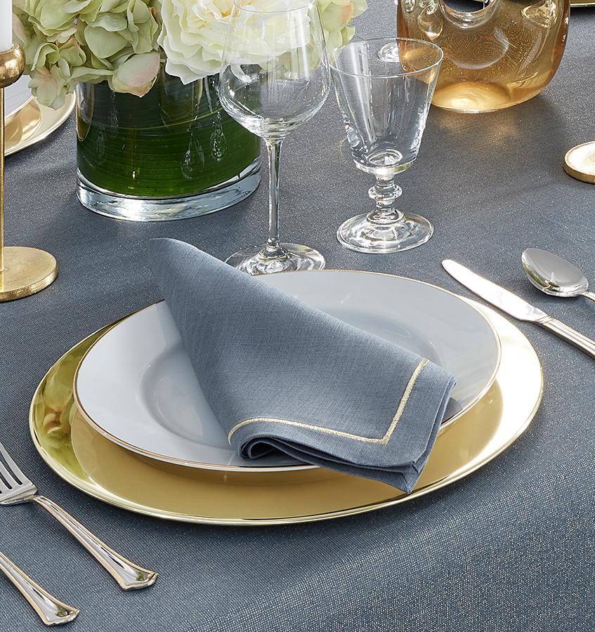 A formal table is set with a white plate with gold rim, a gold charger, and Sferra's Seaton Napkin Flint & Gold Set of 4 atop the plate. Silverware, glassware, and a vase of white flowers sit on a gray tablecloth.