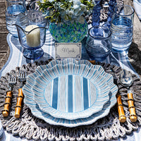 A table setting with Juliska Cabana Stripe Melamine Dinnerware Set of 4 - Ocean, bamboo cutlery, blue glassware, a candle in a blue holder, floral centerpiece, Mark's place card on a woven placemat and striped tablecloth.