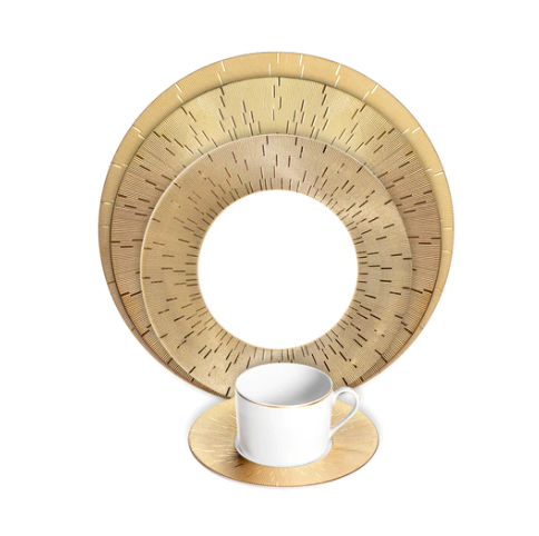 A white teacup with a gold handle rests on a gold saucer, in front of Haviland Infini Prestige Gold Dinnerware, featuring hand-painted Limoges porcelain plates with concentric circular designs and fine lines.