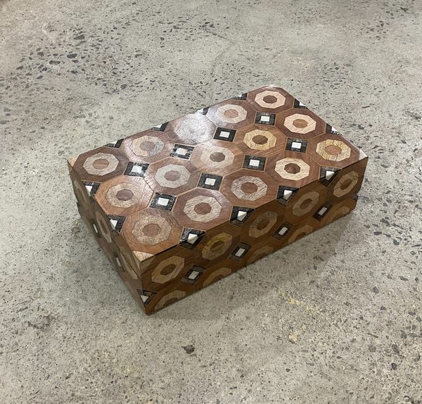 The Bojay Geometric Inlaid Box, featuring an intricate pattern of hexagons and diamonds in brown, beige, and black, sits on a rough gray concrete floor.