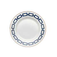 The Catene Zaffiro Dinnerware by Ginori 1735 features a white ceramic plate with a gold rim and bold navy blue chain-link pattern around the edge, inspired by designer porcelain. The center remains plain for an elegant look.