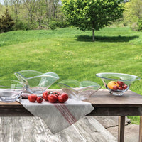 Four Simon Pearce Woodbury Bowls and a red-striped linen towel rest on a rustic wooden table outdoors. Tomatoes are on the towel, with lemons and a tomato in one bowl. Lush green grass and trees form the scenic backdrop.