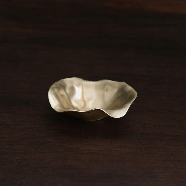 The Beatriz Ball Sierra Maia Oval Gold Bowl, handmade in champagne gold aluminum with wavy edges and a flower-like shape, rests elegantly on dark wood—a stylish, eco-friendly serveware piece.
