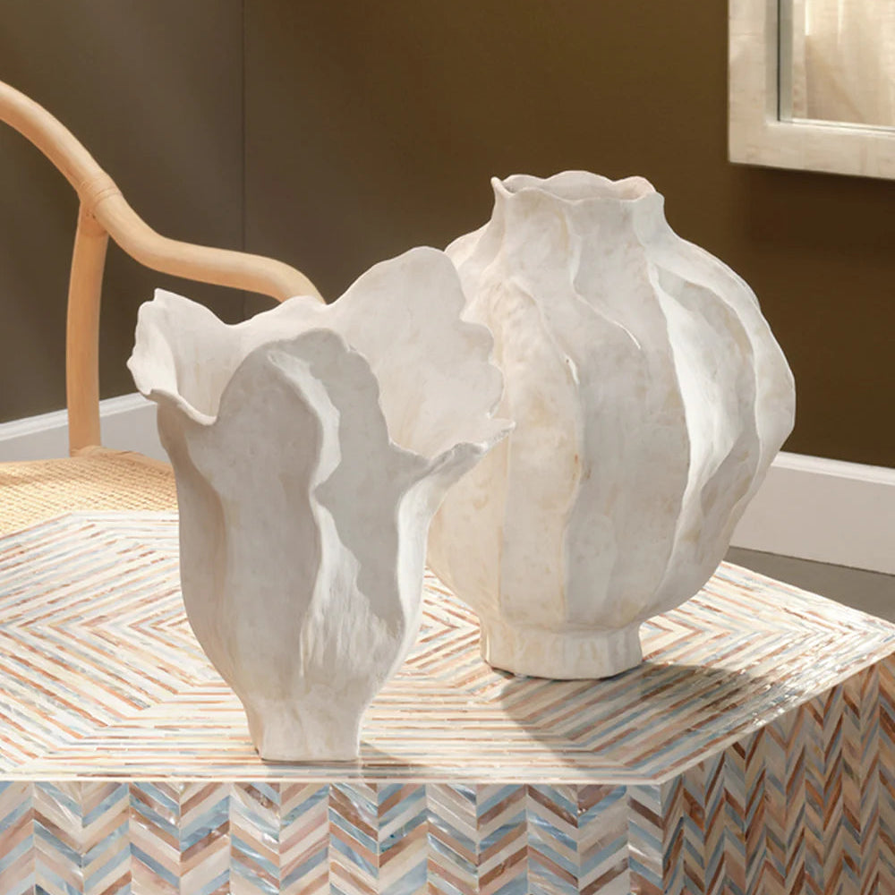 Two white Jamie Young Pompano Vases, artisan-crafted with wavy edges, sit on a patterned table. A light wood chair and brown wall bring warmth to the neutral home decor scene.