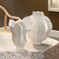 Two white Jamie Young Pompano Vases, artisan-crafted with wavy edges, sit on a patterned table. A light wood chair and brown wall bring warmth to the neutral home decor scene.