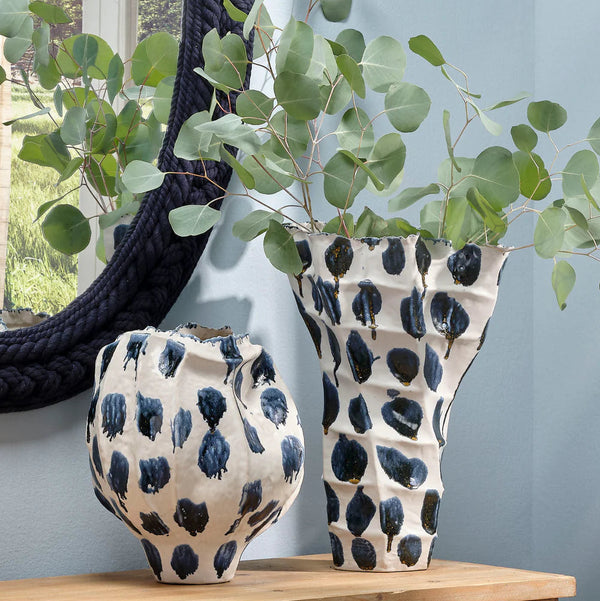 Two Jamie Young Moncler Blue Vases, featuring hand-finished ceramic with blue and black patterns, display eucalyptus branches on a wooden surface near a round navy rope-framed mirror and a window with outdoor greenery.