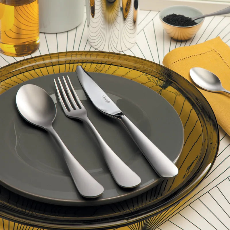A modern table setting features a gray plate on an amber glass plate, complemented by the Christofle Origine Flatware 5 Piece Setting in stainless steel. Also shown: a small bowl of black seasoning, a drink, and a spoon resting on a yellow napkin.