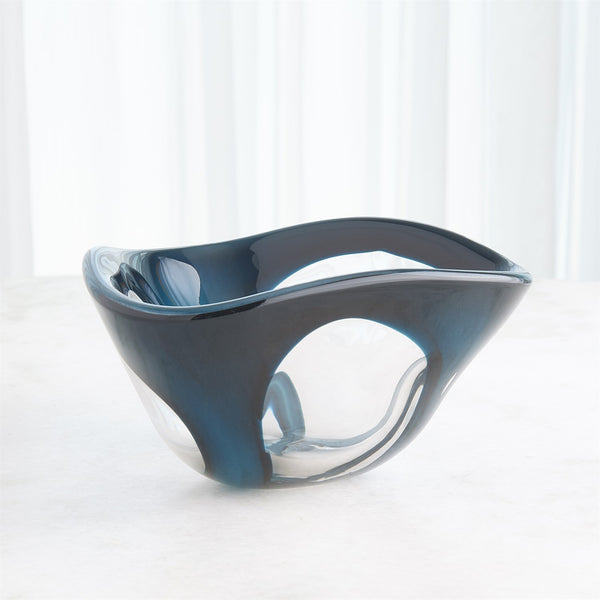 The Enigma Dark Blue Bowl by Global Views / Surya is a handblown art glass piece with a wavy, asymmetrical rim, blue accents, and a unique hollow circular design on one side. It sits on a light surface before sheer white curtains.